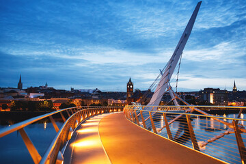 Derry, Ireland. Illuminated Peace bridge in Derry Londonderry, City of Culture, in Northern Ireland with city center at the background. Night cloudy sky with reflection in the river at the dusk