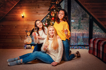 Naklejka premium Christmas and new year. Three young happy women poses with glasses of champagne. Christmas tree on the background. Scandinavian style interior