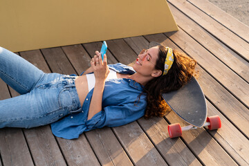 Casual young hipster girl messaging in smartphone using wireless 4g internet connection outdoors lying relaxed in longboard at summer sunset. Trendy dressed stylish female with mobile phone in hands