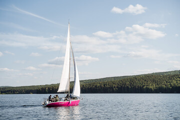 Obraz premium A crimson yacht with raised white sails sails in the middle of the lake. People are resting on the deck.