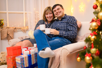 Obraz na płótnie Canvas Man and woman sitting on a couch and posing in new year or christmas decoration. They drinking tea or coffee from white cups.. Holiday lights and gifts, Christmas tree decorated with toys.