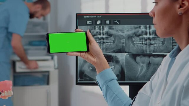 Close up of dentist analyzing smartphone with green screen and teeth scan on computer in dental cabinet. Woman holding device with chroma key for isolated mockup template for dentistry