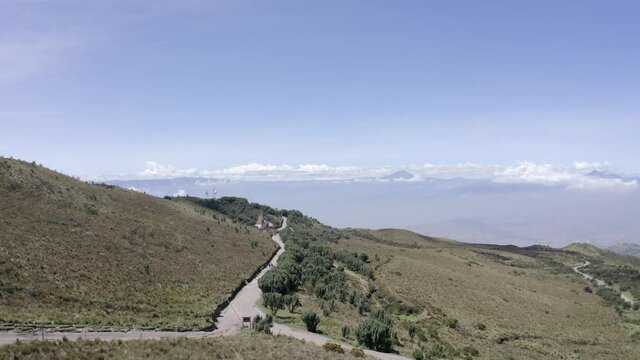 Drone shot of a trail surrounded by trees on the moorlands of the Andes in Rucu Pichincha near Quito, Ecuador. There is a church at the end of the trail. Mountains and a blue sky are seen on the back.