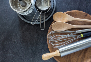 Kitchen utensils, measuring cups, whisk and wooden spoon on dark background with copy space