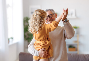 Adorable child girl and positive grandpa holding hands while dancing together in living room