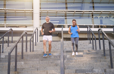 Fototapeta premium Motivated couple, middle aged man and woman in sportswear running up a flight of stairs while training together outdoors in the city