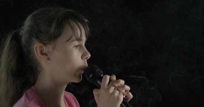 Teenager with a microphone. Cute little girl singing into a microphone on a black background in the smoke.