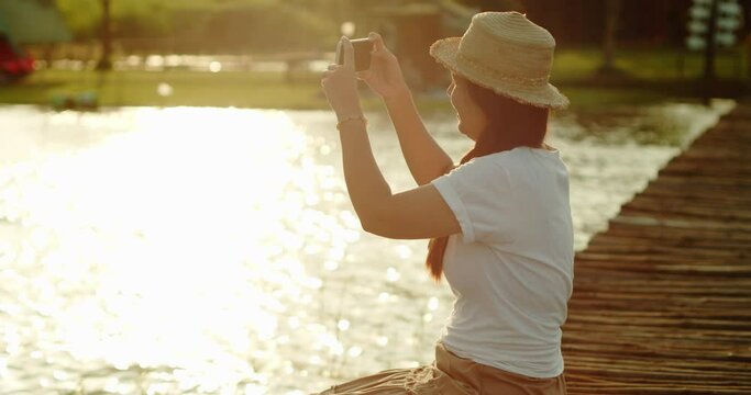 A female tourist in a brown hat, white T-shirt and brown skirt sits on a wooden bridge by the lake, use her mobile phone take the photos relaxing and joyfully in the evening at the sunset time.