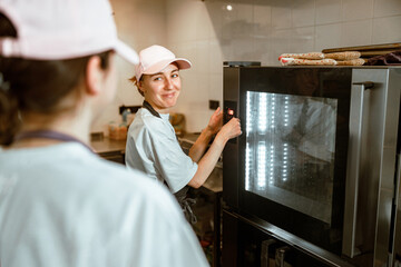 Fototapeta na wymiar Lady in uniform turns on electric oven looking at colleague in modern bakery