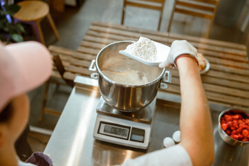 Baker pours white flour into container to weigh on digital scales in workshop