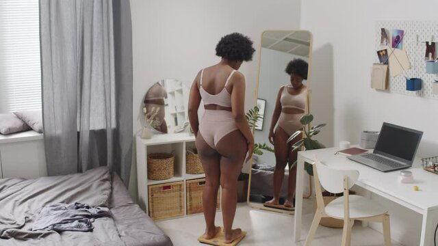 Rear-view full-length slowmo shot of young plus size African-American woman in underwear standing on scales at bedroom and examining her body in mirror