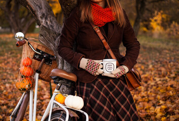 A woman in autumn clothes is drinking tea next to a bicycle on which the halloween pumpkins lie