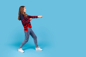 Full length profile portrait of woman wearing striped casual style sweater, standing in attack or pulling hands gesture and looking forward. Indoor studio shot isolated on blue background.