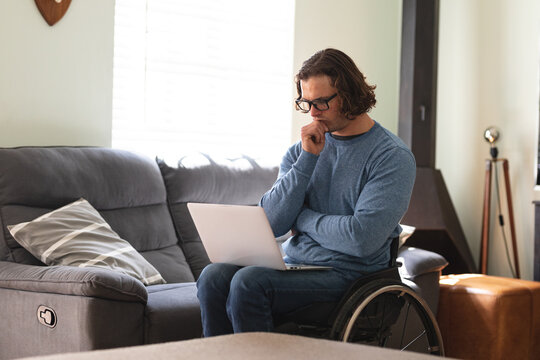 Thoughtful caucasian disabled man wearing glasses sitting on wheelchair using laptop at home