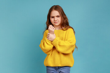 Portrait of little girl standing with grimace of pain, touching sore sprained wrist, health problem, wearing yellow casual style sweater. Indoor studio shot isolated on blue background.