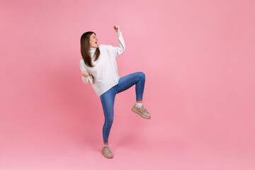 Full length portrait of extremely happy female celebrating her success, standing with clenched fists, wearing white casual style sweater. Indoor studio shot isolated on pink background.