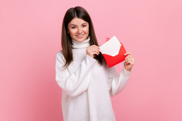 Portrait of woman standing, holding envelope and looking at camera with toothy smile and enjoying, wearing white casual style sweater. Indoor studio shot isolated on pink background. - obrazy, fototapety, plakaty