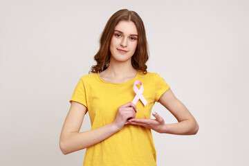 Female health check awareness. Portrait of beautiful teenager girl with brown hair holding pink ribbon in front of chest, support of oncology patients. Indoor studio shot isolated on gray background.