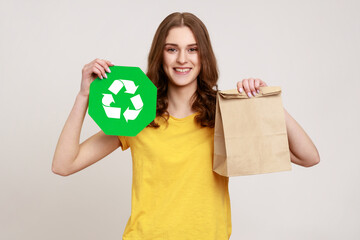 Smiling teenager girl wearing yellow casual T-shirt holding paper package and green recycling sign, looking at camera, expressing happiness. Indoor studio shot isolated on gray background.