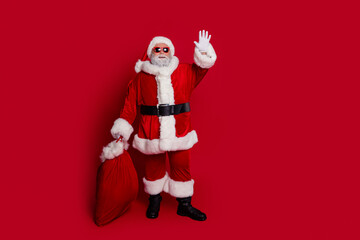 Full body photo of funny santa wave hold bag with presents wear hat eyewear coat pants boots isolated on red background