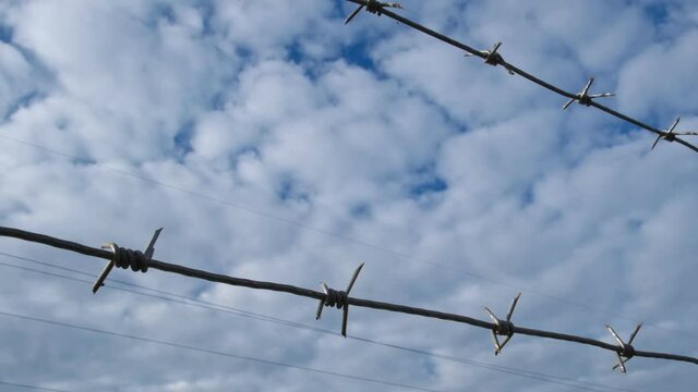 Barbed wire on the background of the cloudy blue sky. Illegal immigration concept, prison, imprisonment, restricted area. Slowly moving along iron fence boundary of the jail. Razor wire fencing.