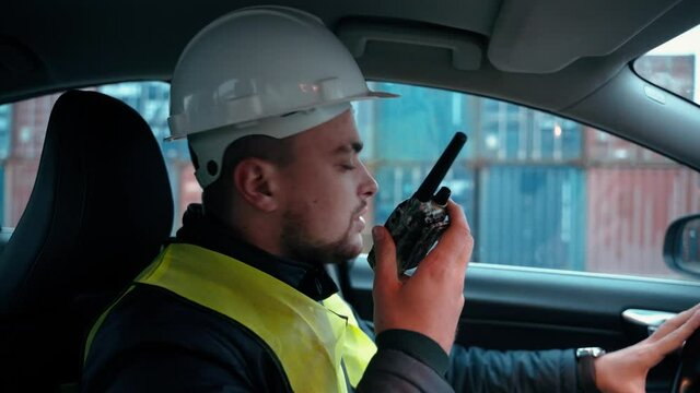 Cargo port worker using radio transmitter while driving his car in containers background. Logistics industry employee talking via portable radio with his colleague
