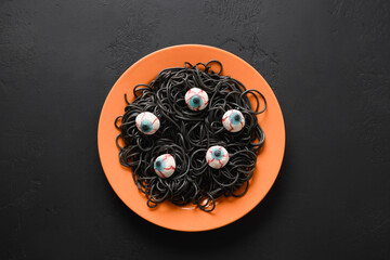 Halloween funny Italian black pasta squid ink decorated horror eyes in orange plate with smoke potion on black background. Horror food for party. View from above.