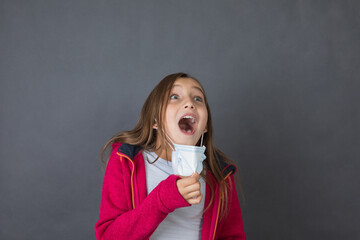 Portrait of a young girl trying to breath with open mouth in red sweater playing with medical mask on grey background. - 463816029
