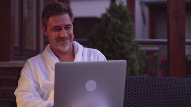Man wearing bathrobe and working on laptop computer in a wellness hotel. Traveling businessman relaxing but working late.