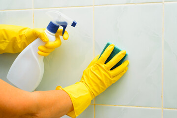 Close-up of female hands in yellow rubber gloves holding sponge and cleaning chemical spray, housewife washing and polishing tiles in bathroom indoors. Weekly disinfection and bath hygiene