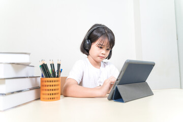 Cheerful Asian little girl student wears headphone, writes on digital tablet to study at home. Education Concept Stock Photo