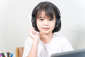 Cheerful Asian little girl student wears headphone, writes on digital tablet to study at home. Education Concept Stock Photo