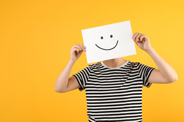 Woman hiding behind sheet of paper with happy face on yellow background, space for text