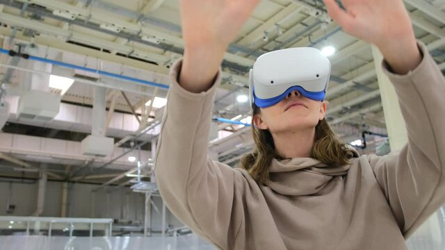 Teenage girl with long dark hair in pink hoodie fumbles with hands having fun with virtual reality glasses in large hall closeup