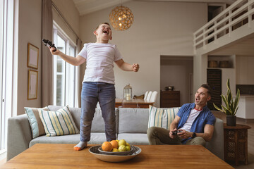 Caucasian boy celebrating standing on the table while playing video games with his father at home