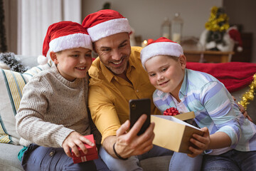 Caucasian father and two sons opening gift box during video call on smartphone during christmas