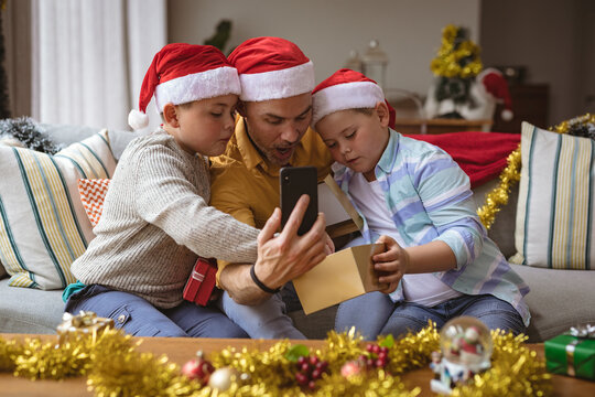Caucasian father and two sons opening gift box during video call on smartphone during christmas