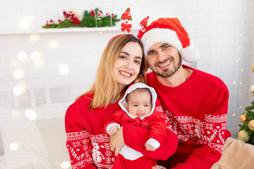 happy young family mom dad and baby in red sweaters under the Christmas tree on the bed at home celebrate New Year or Christmas by kissing and hugging each other