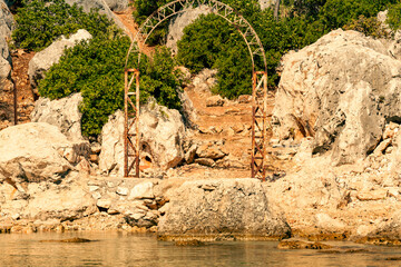 Close-up of the old gate arch and the path leading from the bay of the Mediterranean Sea to the ruins of the old Lycian fortress on the hill. Rocky shore with lush vegetation and sea water. Turkey.