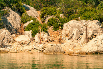 The old gate arch and the path leading from the bay of the Mediterranean Sea to the ruins of the old Lycian fortress on the hill. Rocky shore with lush vegetation and turquoise sea water. Turkey.