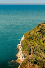 View of a wild sheer cliff overgrown with greenery with the blue water of the mediterranean sea in the background