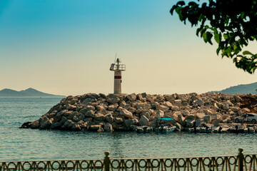 A small lighthouse on a rock ridge in the bay of the town of Kalkan. Mountains in the sea haze on the background. Embankment fence and green trees on the foreground
