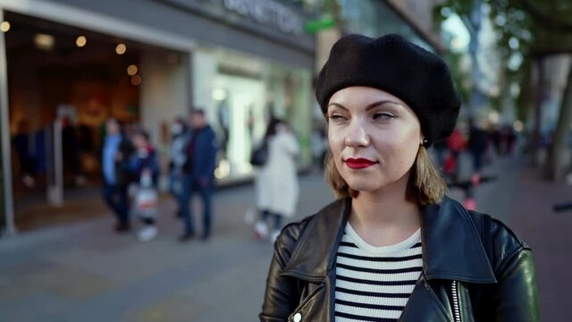 Portrait of trendy european woman in hat standing on busy city street. Lady smiling, modern urban outfit, leather jacket and red lips. 
