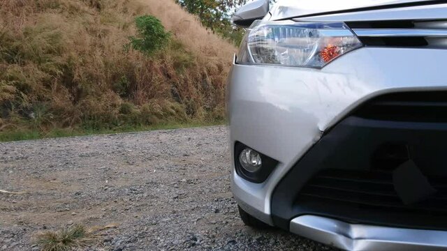 Closeup of silver car front damaged by crash accident with headlight flashing for emergency parking on roadside with gravel road background