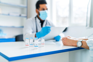 In the focus of the jar with the coronavirus vaccine, on the doctor's desktop vaccine, the doctor consults his patient