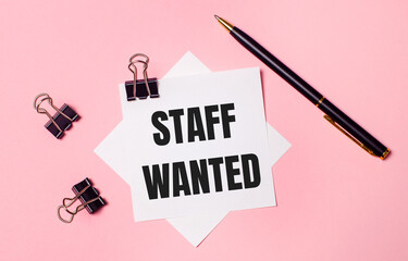 On a light pink background, black paper clips, black pen and white note paper with the words STAFF WANTED. Flat lay