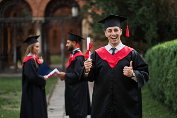 Happy caucasian graduate with his classmates in graduation gown holds diploma in campus