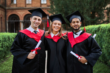 Portrait of three smiling graduate friends in graduation robes in university campus with diploma