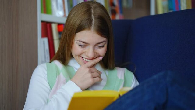 Young woman with long hair wearing casual clothes sitting in armchair and reading funny book, bookshelves on background. Handheld reader enjoying comic literature in library. Concept of hobby