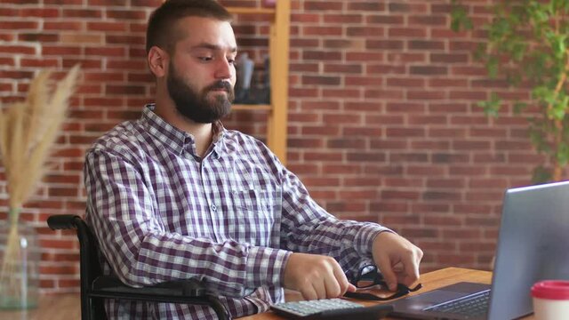 Tired hipster businessman is sitting on wheelchair at table with laptop and rubbing his eyes. Disabled person works remotely. Concept of freelancing and remote work for people with disabilities